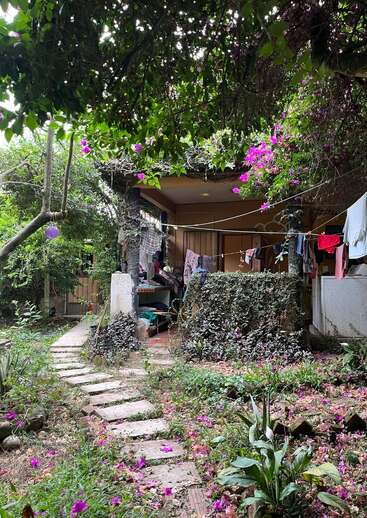 The image depicts a serene outdoor scene featuring a house with a clothesline, surrounded by lush greenery and vibrant purple flowers, with a stone pathway leading to the entrance.