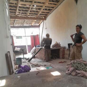 L'image représente une pièce en cours de rénovation, avec deux personnes debout au milieu d'outils et de matériaux épars, entourées d'un espace encombré de poutres apparentes et d'un toit partiellement ouvert.