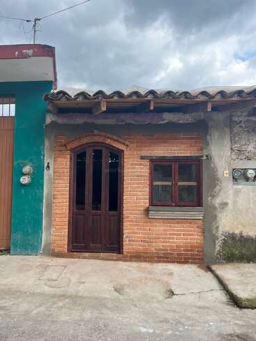 Cette image montre une petite maison en briques avec une porte et une fenêtre en bois, un toit en tuiles, des murs non finis, des bâtiments adjacents, un trottoir en béton et un ciel nuageux.
