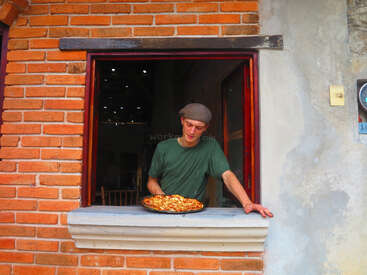 A man wearing a green shirt and a flat cap leans out a brick window, presenting a large pizza on a tray with a proud expression.