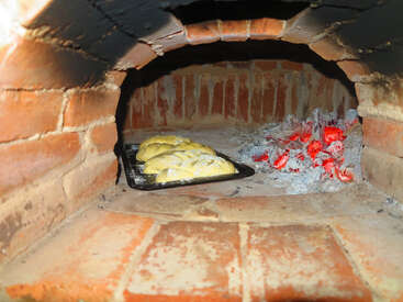 Inside a rustic brick oven, dough rises on a baking tray next to glowing red-hot coals, creating a warm, traditional atmosphere of homemade bread baking.