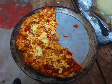 A partially eaten cheese and tomato pizza sits on a round, well-used metal baking tray. Nearby are kitchen utensils and some dough on a floured surface.