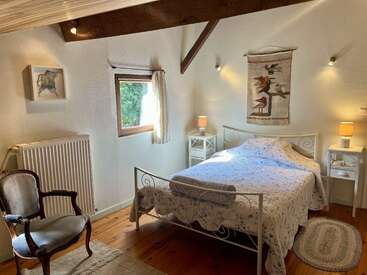 Um quarto aconchegante apresenta uma cama de casal com colcha estampada, pisos de madeira, iluminação suave, vigas rústicas, uma poltrona elegante e decorações artísticas nas paredes.