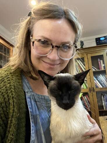 Eine Frau mit Brille, die einen grünen Pullover trägt, lächelt, während sie eine Siamkatze hält. Im gemütlichen Hintergrund sind Bücherregale und Holzmöbel zu sehen.