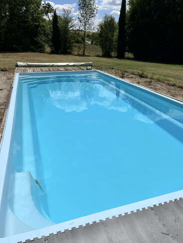 Uma piscina retangular azul clara fica em um quintal gramado, cercada por árvores e vegetação, refletindo o céu e as nuvens, criando uma cena tranquila ao ar livre.