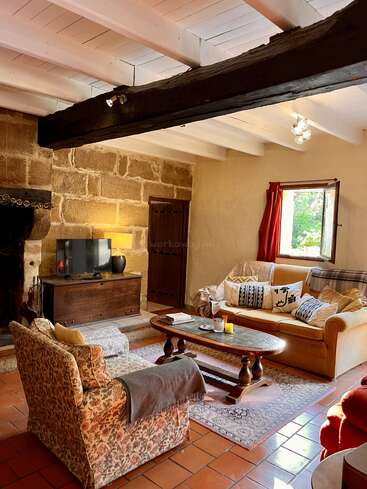 Uma sala de estar aconchegante com paredes rústicas de pedra, vigas de madeira, poltronas confortáveis, um sofá amarelo, mesa de centro, TV, iluminação aconchegante e uma janela aberta com luz solar.