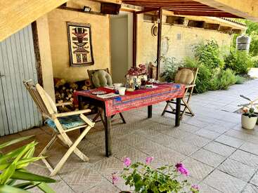 Um charmoso pátio externo apresenta uma mesa com um pano colorido, duas cadeiras de madeira, vasos de plantas, decoração rústica e a luz do sol iluminando um ambiente aconchegante e convidativo.