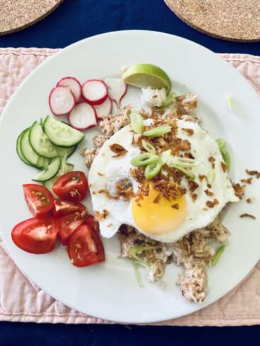 Um prato com arroz frito coberto com um ovo virado para o sol, guarnecido com cebolinha verde e chalotas crocantes, rodeado por pepino fresco, rabanete, tomate e limão.