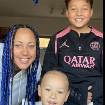 Une femme souriante aux tresses bleues pose à l'intérieur avec deux enfants ravis. L'aîné porte une veste noire du Paris Saint-Germain. Un moment familial chaleureux et joyeux.
