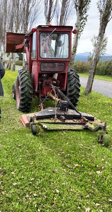 Um trator vermelho com pneus grandes e um cortador de grama traseiro está sobre a grama verde, cercado por árvores, céu nublado e uma estrada rural próxima.