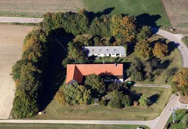 Das Bild zeigt eine ländliche Landschaft mit einem großen Haus und Nebengebäuden, mit einer langen Auffahrt und einem Feld, umgeben von Bäumen und einem Zaun, mit einem klaren Himmel darüber.