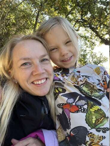 A woman and young girl smile for a close-up selfie outdoors. The girl wears a butterfly-patterned shirt. Sunlight filters through trees, creating a joyful and warm atmosphere.