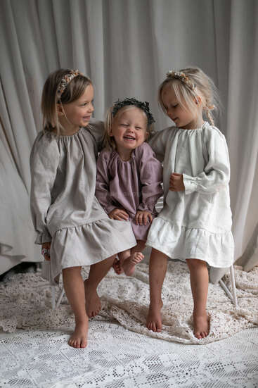 Three young girls sit together on a lacy blanket, smiling joyfully. They wear pretty, neutral-toned dresses and decorative headbands, radiating warmth and innocence. Curtains backdrop.
