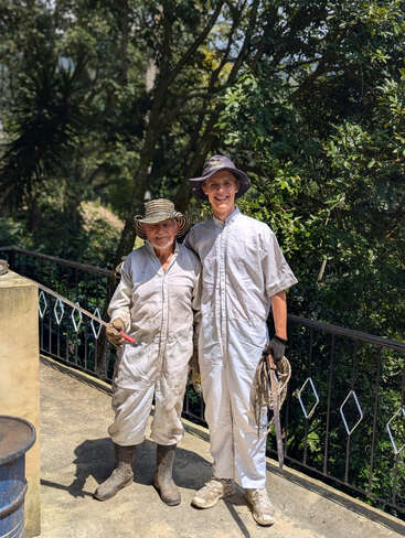 Dos hombres vestidos con monos blancos, gorras y botas están de pie al aire libre en un día soleado, sonriendo. Parecen ser trabajadores agrícolas, rodeados de una exuberante vegetación.