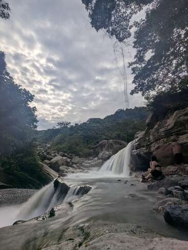 Una tranquila cascada cae sobre las rocas y desemboca en un estanque apacible. Una exuberante vegetación rodea la escena. Dos personas se relajan sobre las rocas, disfrutando de la tranquila belleza de la naturaleza.