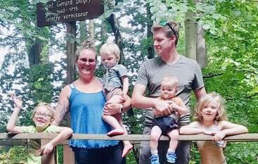 A family of two adults and four children pose together outdoors on a wooden bridge, surrounded by green trees, smiling and enjoying a sunny day in nature.