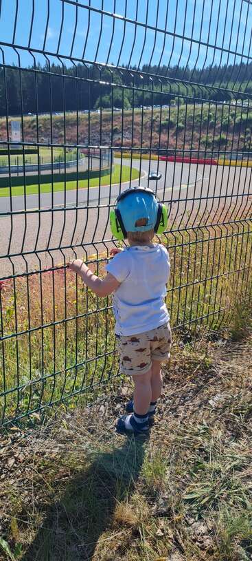 A young child stands behind a fence at a racetrack, wearing a blue cap and green earmuffs, watching the cars race under a sunny sky.