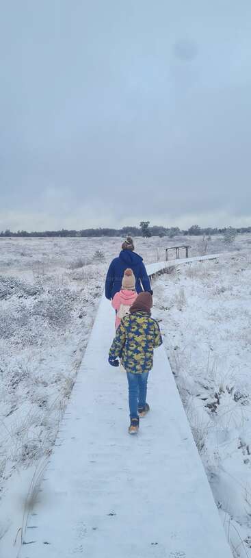 A woman and two children, warmly dressed, walk along a snowy path through a wintery landscape, surrounded by frosty fields under a cloudy sky.