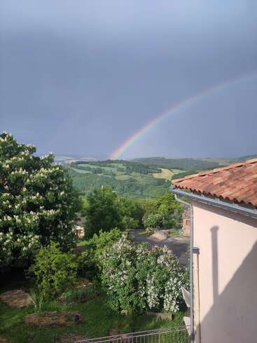 A vibrant rainbow arches across a cloudy sky over lush green hills, blooming trees, and a tiled-roof house, creating a peaceful countryside landscape after rain.