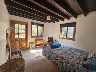This cozy bedroom features a floral bed, exposed dark ceiling beams, wooden furniture, large windows with blinds, a wicker basket, and a sunlit door.