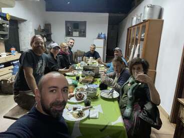 A group of ten people are gathered around a dining table, enjoying food and drinks together in a cozy, rustic room. Some are smiling.