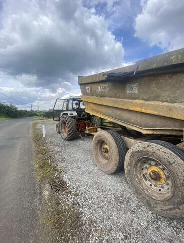 A tractor with a large attached trailer is parked on a gravel roadside under a cloudy sky. The scene suggests rural or agricultural activity and a country road.