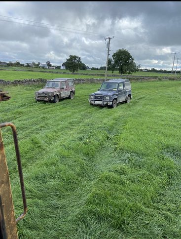 Two muddy off-road vehicles are parked on lush green grass under a cloudy sky. Stone walls and power lines border open countryside with distant houses.