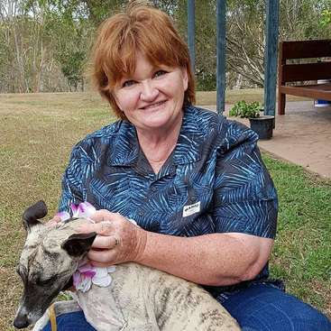 The image depicts a woman sitting on the grass, cradling a dog adorned with a flower collar, set against a serene outdoor backdrop featuring trees and a bench.