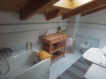 This cozy attic bathroom features a bathtub, wooden shelving, bidet, sink, and skylight. White tiles, wooden beams, and a striped rug create a warm, inviting atmosphere.