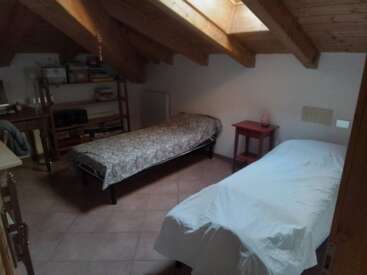 This cozy attic bedroom features two single beds, a wooden ceiling with skylights, a small red nightstand, a desk, shelves, and tiled flooring.