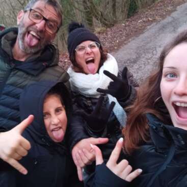 Four people pose happily outdoors, making silly faces and hand gestures. They all stick out their tongues, dressed warmly in jackets, hats, and scarves, enjoying themselves.