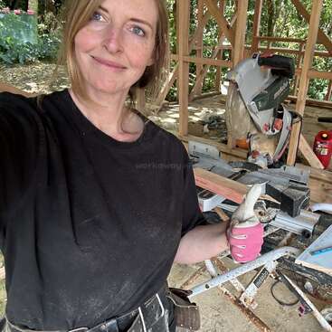 Une femme portant une chemise noire et un gant de travail rose sourit, levant le pouce sur un chantier de construction avec du bois et une scie circulaire derrière elle.