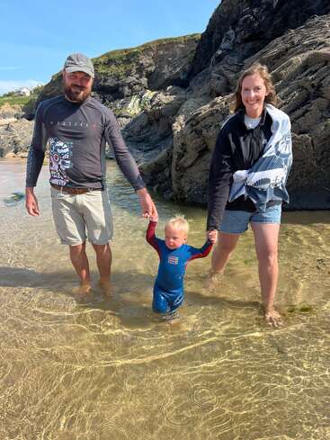 Un homme et une femme souriants tiennent la main d'un petit enfant, marchant ensemble dans l'eau peu profonde de la plage, entourée de falaises rocheuses sous un ciel bleu lumineux.