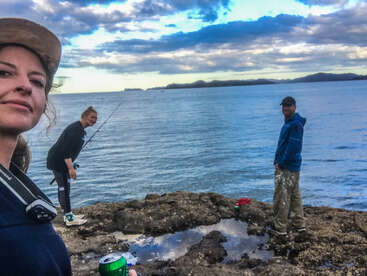 Trois personnes profitent d'une journée au bord de l'eau sur un rivage rocheux : elles pêchent, se détendent et prennent des selfies avec des boissons, sous un ciel bleu partiellement nuageux.
