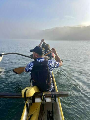 Trois personnes pagaient dans un canoë à balancier jaune sur une eau calme, entourée de brume. La lumière du soleil éclaire doucement la scène, évoquant une atmosphère sereine et aventureuse.