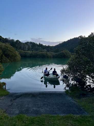 Deux personnes dans un petit bateau pagaient sur un lac paisible, d'un bleu verdâtre, entouré d'arbres, le soleil se couchant derrière de lointaines collines boisées. Atmosphère paisible en soirée.