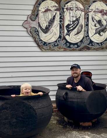 Un jeune enfant et un homme adulte sont assis en jouant à l'intérieur de deux grands chaudrons noirs, souriants, avec en arrière-plan des œuvres d'art sur le thème du nautisme et un revêtement gris.
