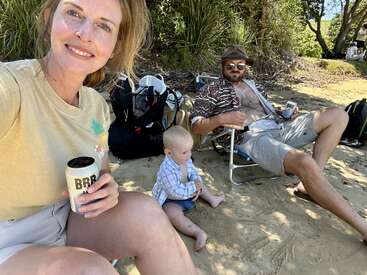 Une famille se détend sur une plage de sable. Une femme prend un selfie en tenant une boisson. Un homme se prélasse à côté d'un bébé qui joue dans le sable.