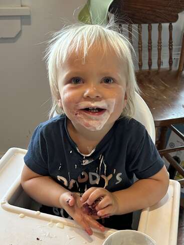 Un enfant souriant aux cheveux blonds en désordre est assis dans une chaise haute, le visage et la chemise couverts de yaourt, jouant joyeusement avec la nourriture, profitant d'un moment joyeux à l'heure du repas.