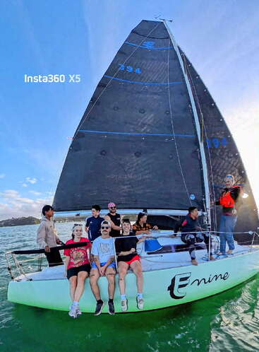 Un groupe de neuf personnes profite d'une journée ensoleillée en naviguant sur un bateau blanc nommé "Enine", se détendant et souriant sous le vaste ciel bleu et la voile sombre.