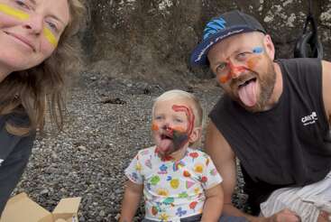 Eine dreiköpfige Familie mit bunt geschminkten Gesichtern genießt die Zeit im Freien an einem felsigen Strand. Die Erwachsenen und das Kind lächeln und strecken spielerisch ihre Zungen heraus.