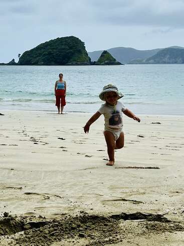 Ein Kind mit Hut rennt fröhlich über einen Sandstrand, während ein Erwachsener am Wasser steht. Im Hintergrund sind grüne Inseln und Hügel zu sehen.