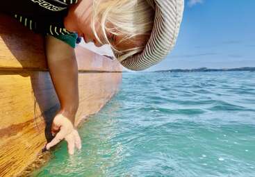 Ein Kind mit einem gestreiften Hut lehnt sich über den Rand eines Holzbootes und berührt sanft das klare blaue Wasser an einem sonnigen Tag.