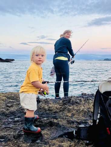 Ein kleines Kind in Gummistiefeln steht am felsigen Ufer und hält eine Angelrute. Ein Erwachsener angelt in der Nähe, mit dem Meer und dem bewölkten Himmel im Rücken.