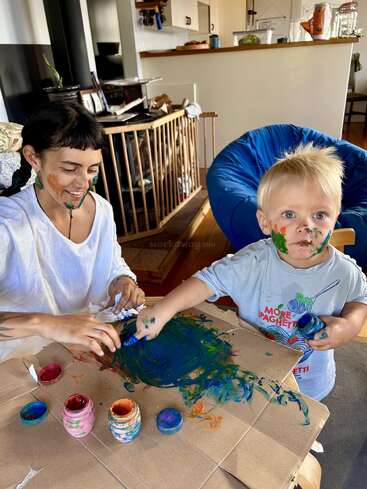 Una mujer y un niño pequeño disfrutan juntos pintando de forma desordenada en una mesa. Ambos tienen pintura de colores en la cara y sonríen, creando arte en un acogedor entorno doméstico.