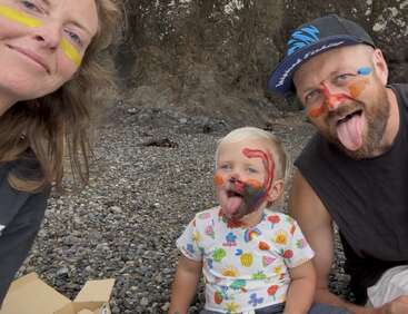A family of three sits on a rocky beach, their faces painted colorfully. The child and father stick out their tongues, smiling happily, enjoying playful fun.