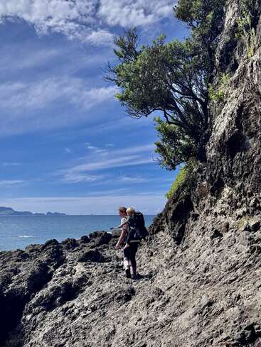Une personne portant un enfant dans un sac à dos marche le long d'une côte rocheuse, sous un ciel bleu parsemé de nuages, au milieu des arbres et des montagnes lointaines qui se fondent dans la mer calme.