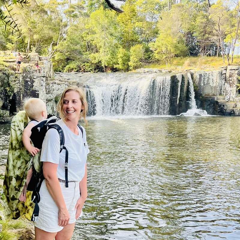 Une femme souriante portant un bébé dans un sac à dos se tient près d'une cascade sereine, entourée d'arbres verdoyants et d'une eau claire et ondulante.