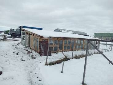 A small, rustic structure with many windows sits in a snowy landscape. Nearby fencing and vehicles suggest it’s a farm or rural area under overcast skies.