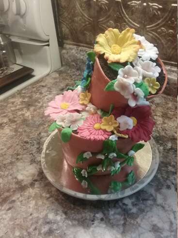 This is a whimsical, tilted cake designed to look like flower pots overflowing with colorful fondant flowers and leaves, sitting on a marble countertop.